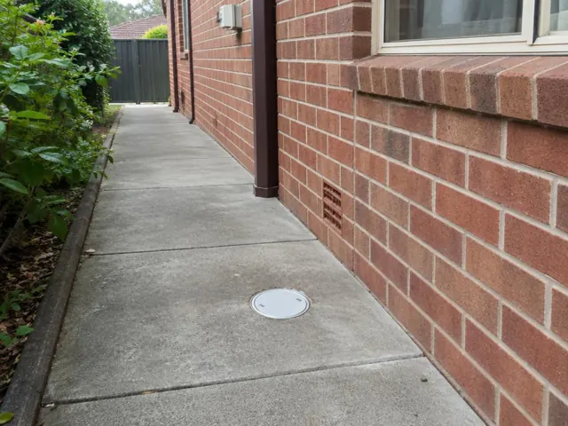 Concrete side path beside a brick house with a round drain cover and dry paving in Thornleigh