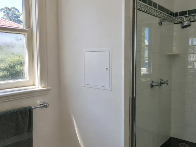 Small access panel in a painted bathroom wall beside a tiled shower and window in Normanhurst